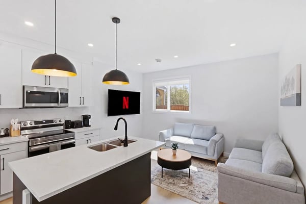 Modern open-plan kitchen and living room with white cabinetry, stainless steel appliances, black pendant lights, and gray seating area with windows.