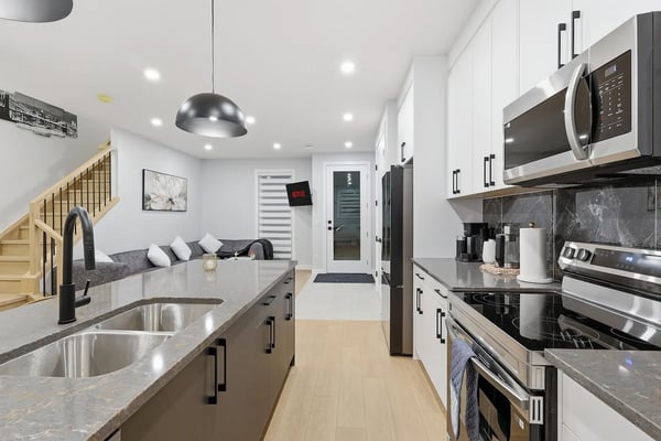 Modern galley kitchen with white cabinetry, stainless steel appliances, gray countertops, and wood flooring opening to living area.